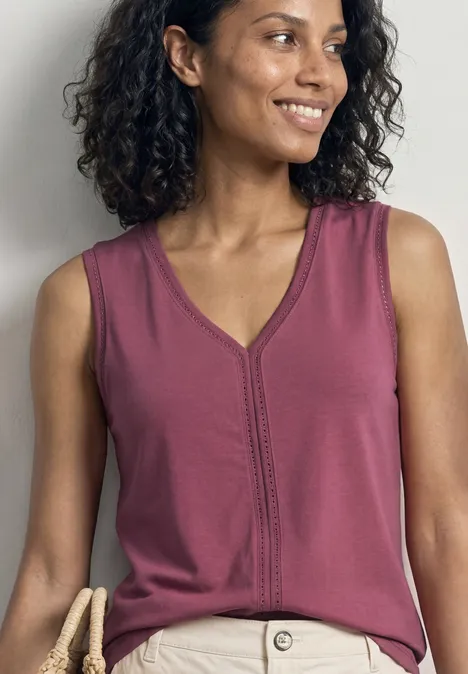 Frau mit lockigem Haar in ärmellosem, violettem Top mit V-Ausschnitt, vor hellem Hintergrund.