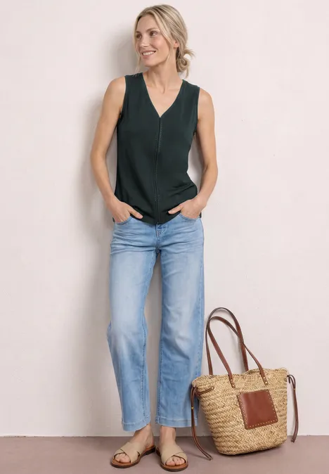 Frau in dunkelgrünem ärmellosem Top, blauen Jeans, Sandalen und Strohtasche vor neutralem Hintergrund.