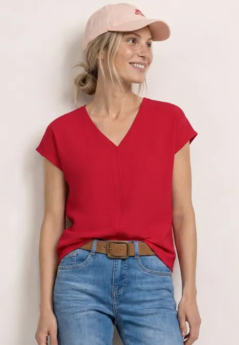 Frau in roter V-Bluse, Jeans und rosa Basecap, vor weißer Wand