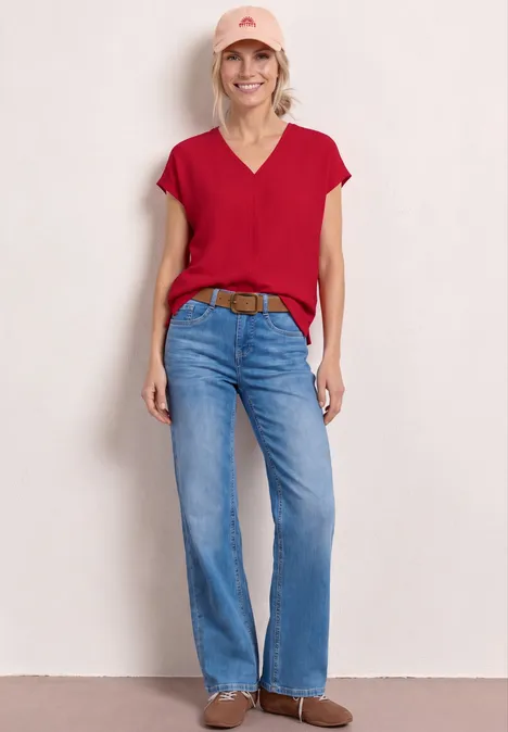 Frau in roter Bluse, blauer Jeans, braunem Gürtel und Stiefeletten vor hellem Hintergrund.