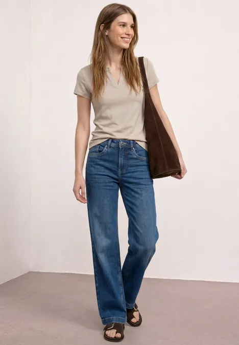 Junge Frau mit T-Shirt, Jeans, Sandalen und Tasche vor neutralem Hintergrund.