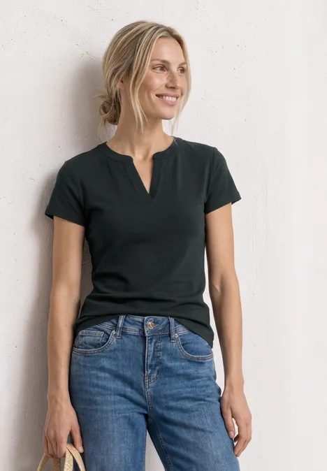 Frau in schwarzem T-Shirt und blauen Jeans lehnt entspannt an weißer Wand.