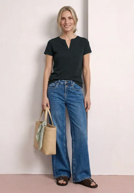 Frau in schwarzem T-Shirt, weiten Jeans, Sandalen, trägt eine geflochtene Tasche mit Tuch.