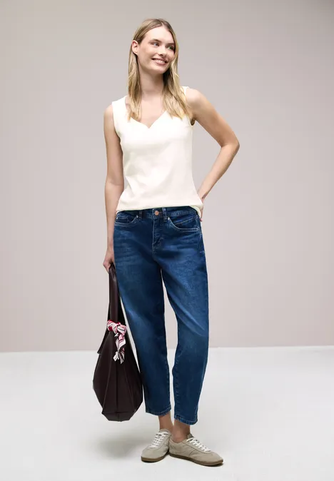 Frau in weißem Top, blauen Jeans und beigen Schuhen hält eine dunkelviolette Tasche mit Tuch, vor hellem Hintergrund.