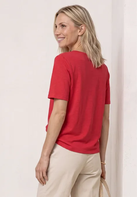 Iemand staat met de rug naar de camera, draagt een rood T-shirt en een beige broek tegen een lichte achtergrond.