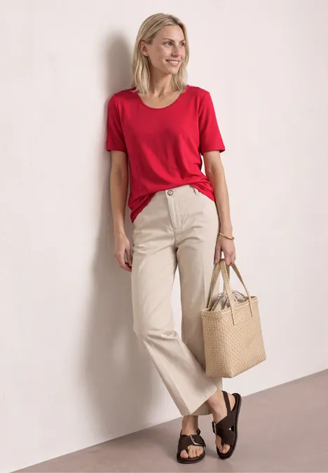 Vrouw staat voor witte muur, draagt rood T-shirt, lichtgekleurde broek, bruine sandalen en een beige tas.