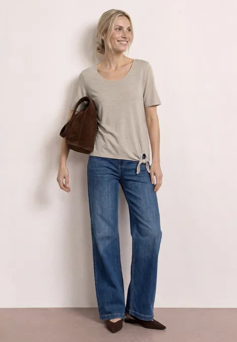 Een persoon draagt een grijs t-shirt, wijde blauwe jeans, zwarte schoenen en een bruine tas.