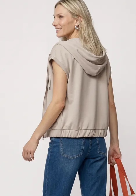 Frau trägt ärmellose Kapuzenjacke und Jeans, hält eine orangefarbene Tasche in der Hand
