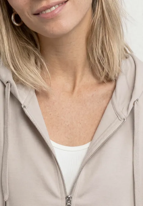 Halbportrait einer lächelnden Frau in beigem Kapuzenpullover.
