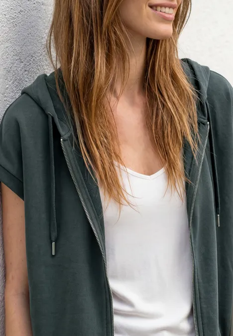 Vrouw in donkergroene mouwloze hoodie over wit topje, lacht en kijkt opzij.