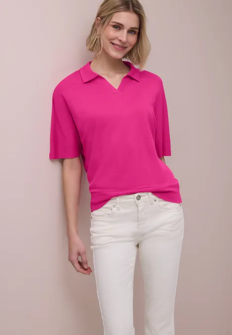 Frau mit pinkem Poloshirt und weißen Jeans vor hellbeigem Hintergrund.