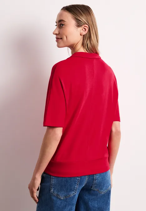 Poloshirt met elastische tailleband salsa red Een vrouw met een rood T-shirt en blauwe spijkerbroek, gezien van achteren tegen een lichte achtergrond.