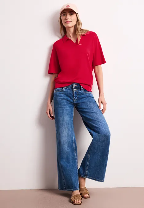 Poloshirt met elastische tailleband salsa red Vrouw met rood T-shirt, wijde blauwe jeans en sandalen, leunend tegen een muur.