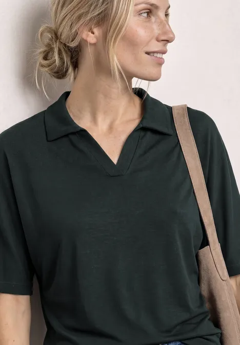 Poloshirt met elastische tailleband deep bound green Vrouw in een donker poloshirt, haar in een losse knot, draagt een stoffen tas over haar schouder.