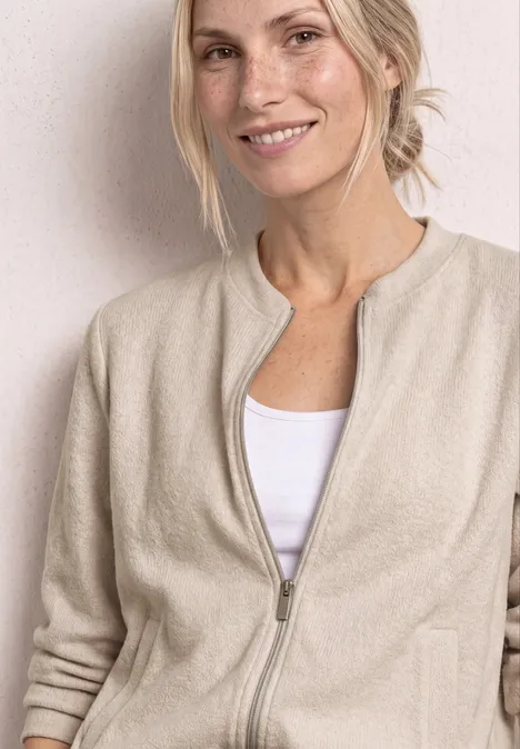 Eine lächelnde Frau im beigen Reißverschluss-Pullover und weißem Top vor hellem Hintergrund