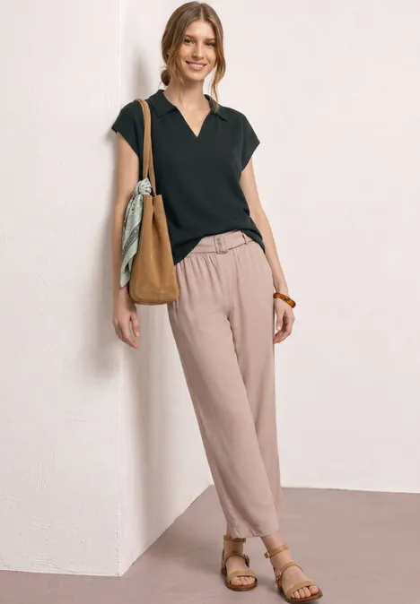 Vrouw leunt nonchalant tegen muur, zwarte V-top, lichte losse broek, sandalen en schoudertas.