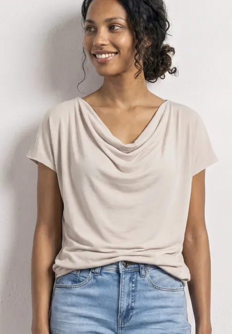 Een persoon draagt een crèmekleurig drapeeshirt met korte mouwen en een blauwe spijkerbroek op een lichte achtergrond.