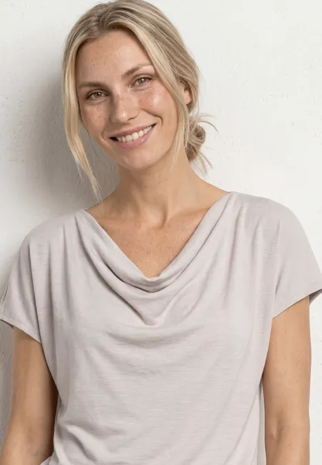 Helle Person mit blondem Haar in lockerem Shirt mit Wasserfallausschnitt vor schlichter weißer Wand.