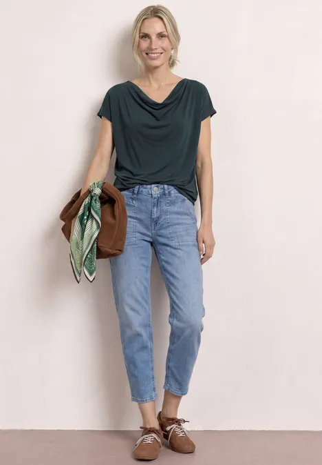 Shirt met hangende schouders en watervalvoorkant deep bound green Vrouw met groen shirt, lichtblauwe high waist jeans en bruine loafers, houdt jas en sjaal vast.