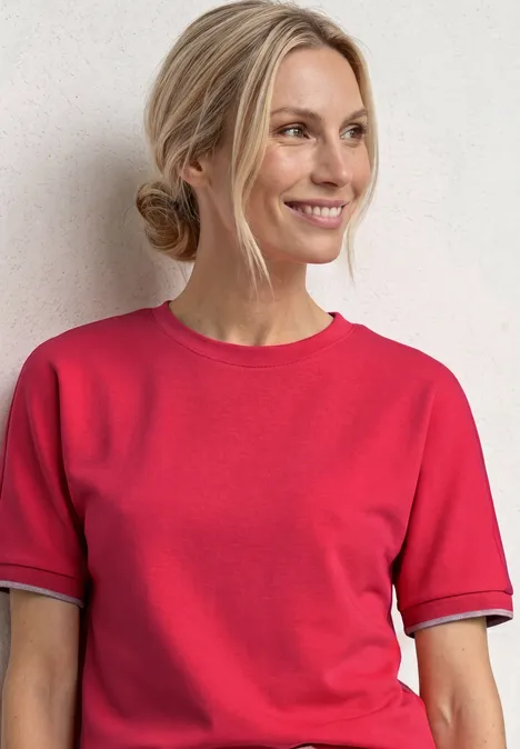 Frau mit hochgestecktem Haar trägt rotes T-Shirt vor hellem Hintergrund.