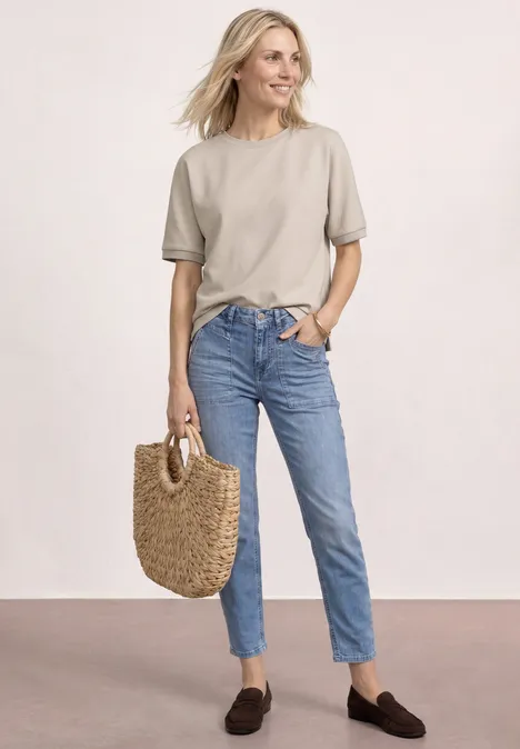 Vrouw in beige T-shirt, blauwe jeans en bruine loafers met een strooien tas, casual uitstraling.