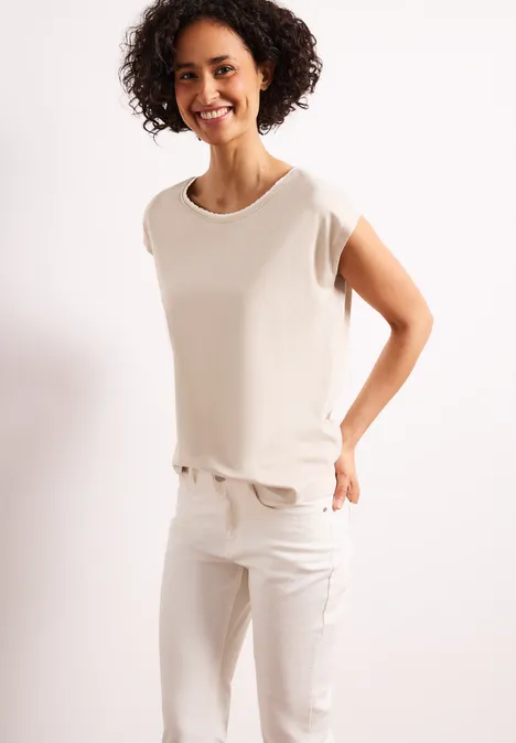 Vrouw in wit t-shirt en witte broek poseert voor een lichte achtergrond.