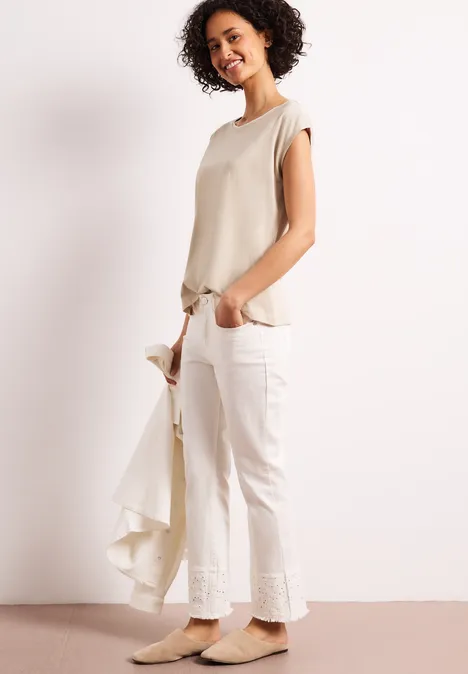 Vrouw met beige top en witte broek, houdt licht jasje vast tegen een witte achtergrond.