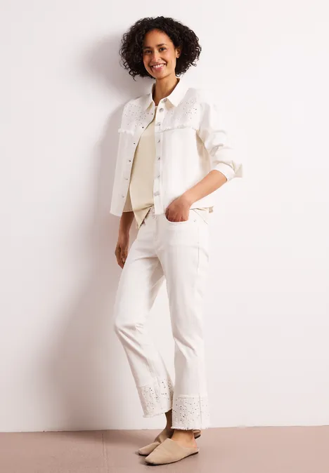 Vrouw met krullend haar draagt witte spijkerjas en broek, beige platte schoenen, leunend tegen een lichte muur.