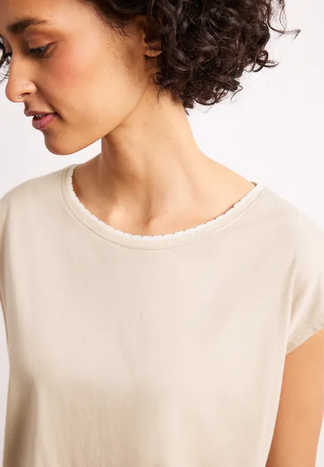 Persoon met donker krullend haar draagt crème T-shirt met ronde hals.