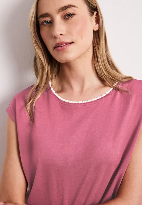 Une personne porte un haut sans manches rose avec un col blanc, debout devant un mur blanc.