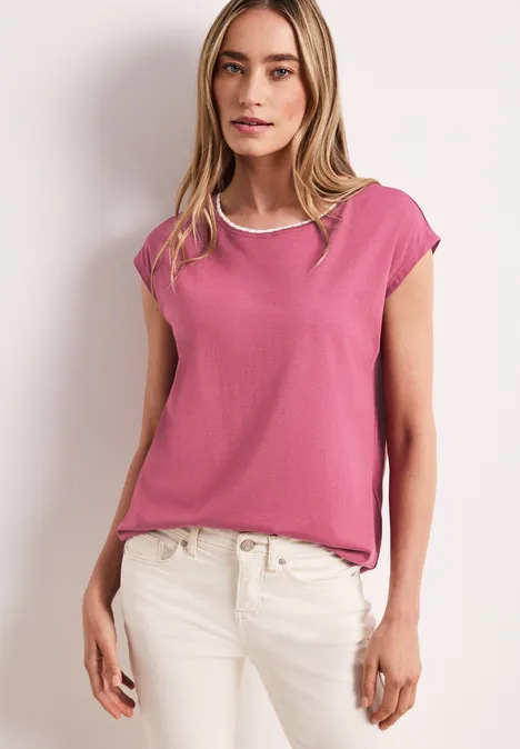 Femme portant un t-shirt rose et un jean blanc devant un mur gris clair.