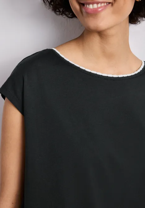 Shirt met overhangende schouder en decoratieve tape deep bound green Een glimlachende vrouw draagt een eenvoudig zwart shirt met witte ronde hals, haar krullen omlijsten het gezicht.