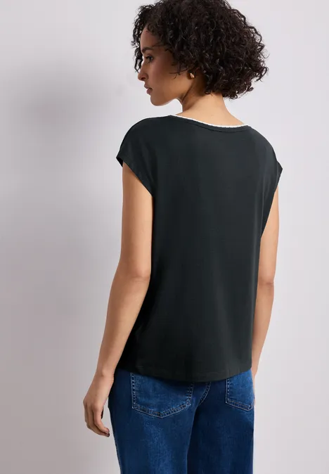 Shirt met overhangende schouder en decoratieve tape deep bound green Vrouw van achteren in een los zwart T-shirt en een blauwe spijkerbroek tegen een lichte achtergrond.