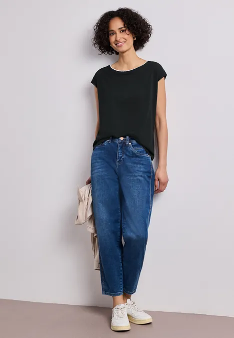 Eine Frau mit lockigem Haar trägt schwarzes Top, blaue Mom-Jeans, weiße Sneaker und hält eine beige Jacke.