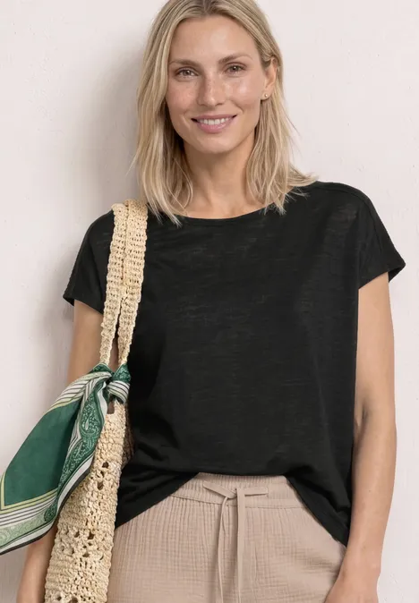 Frau mit schwarzem T-Shirt, beiger Hose und geflochtener Schultertasche mit grünem Tuch.