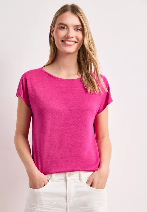 Frau steht mit Händen in den Taschen, trägt pinkes T-Shirt und weiße Hose vor hellem Hintergrund.