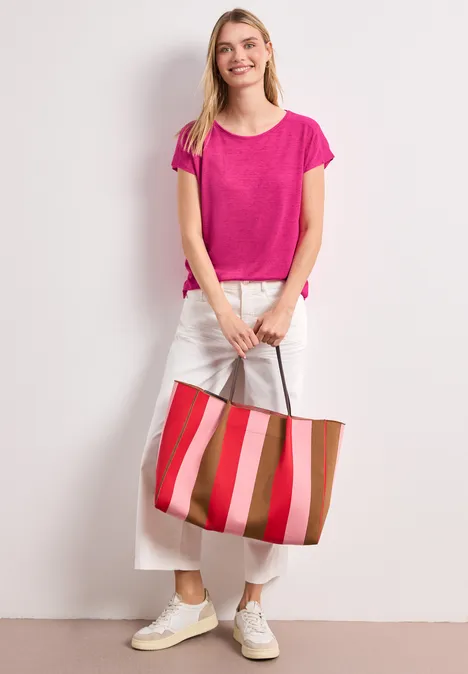 Person steht vor heller Wand, trägt pinkes Shirt, weiße Hose, weiße Sneaker und hält eine gestreifte Tote-Bag.