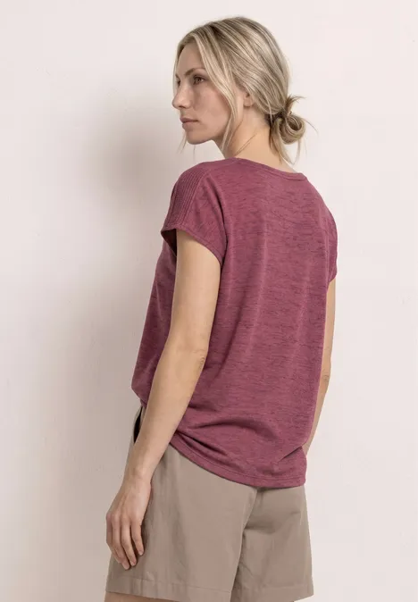 Frau steht in weinrotem T-Shirt und beigen Shorts, Rückenansicht, Blick abgewandt.