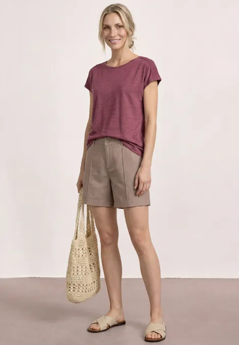 Frau in lässigem Sommerlook mit bordeauxfarbenem T-Shirt, beigen Shorts, geflochtener Tasche und Sandalen.