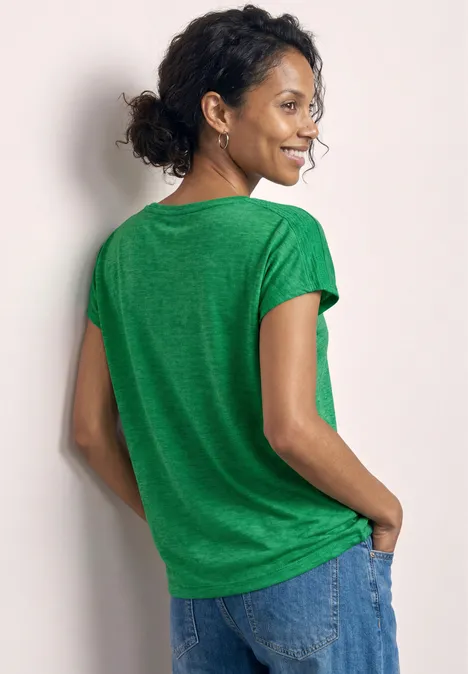 Eine Person steht mit dem Rücken zur Kamera, trägt ein grünes T-Shirt und Jeans, Hände in den Taschen.