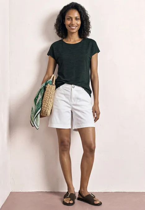 Frau in schwarzem Shirt, weißen Shorts und Sandalen mit Strohtasche und buntem Tuch.