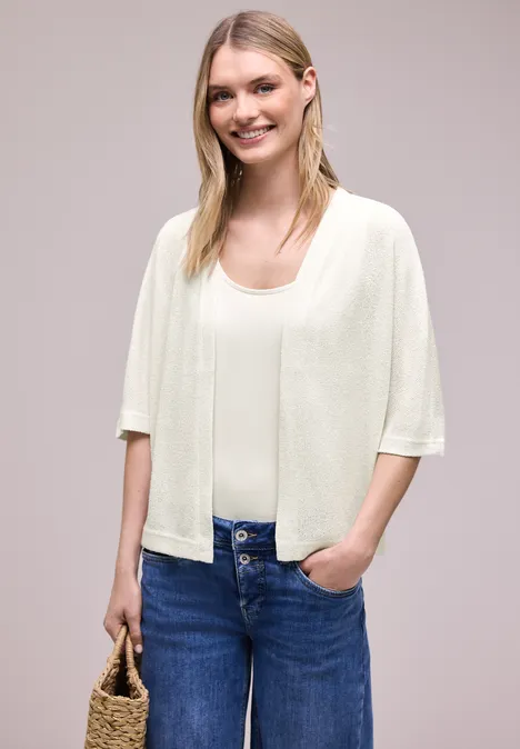 Frau trägt cremefarbenes Kurzarm-Cardigan über Top, blaue Jeans und hält eine Strohtasche.