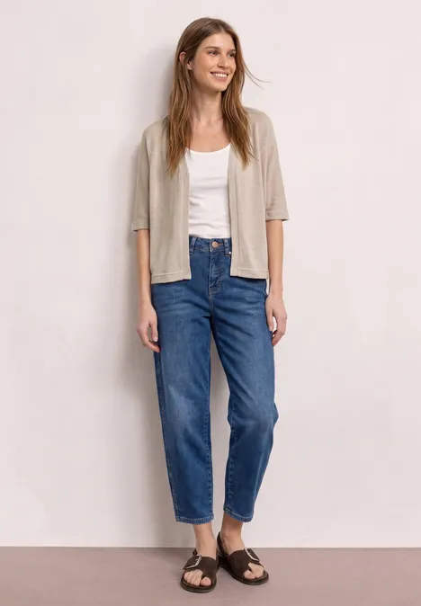 Frau mit unscharfem Gesicht vor neutralem Hintergrund in weißem Top, beigem Cardigan, blauen Jeans und braunen Sandalen.