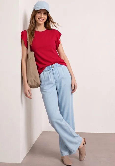 Eine Frau lehnt an der Wand, trägt rotes T-Shirt, helle Jeans und eine Umhängetasche.