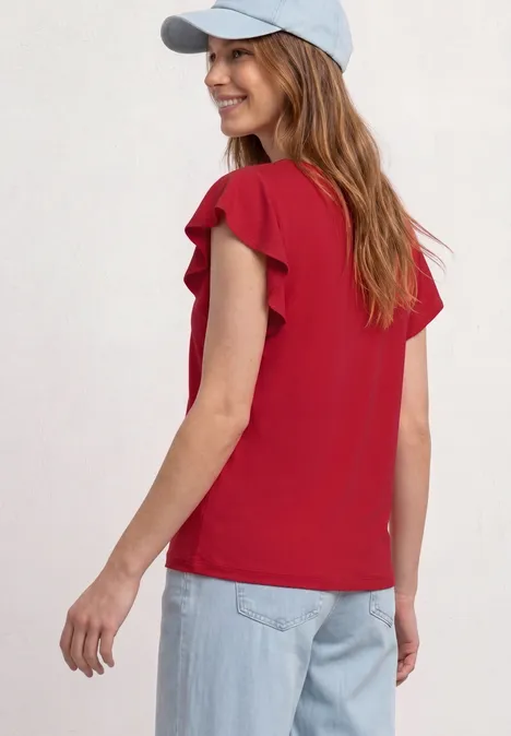 Frau steht vor hellem Hintergrund, trägt rote, kurzärmlige Bluse mit Volants, blaue Jeans und weiße Kappe.