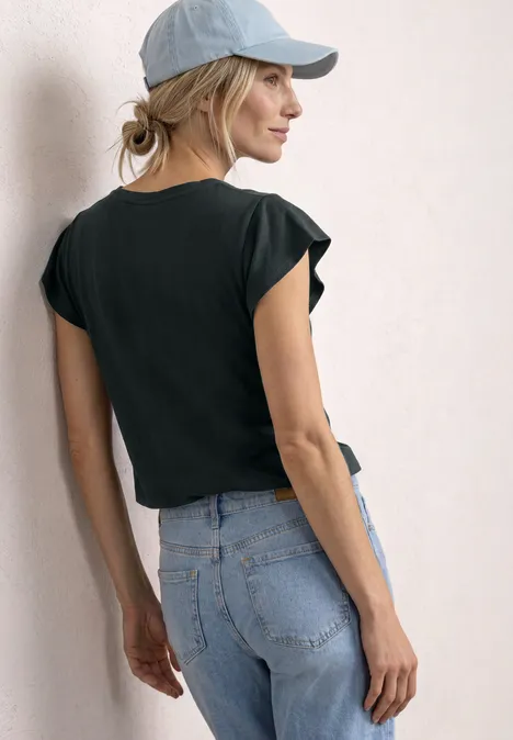 Vrouw van achteren in zwart T-shirt, lichte jeans en pet voor een witte muur