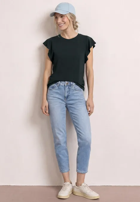 Vrouw in zwart T-shirt, lichtblauwe spijkerbroek, witte sneakers en pet voor neutrale achtergrond.