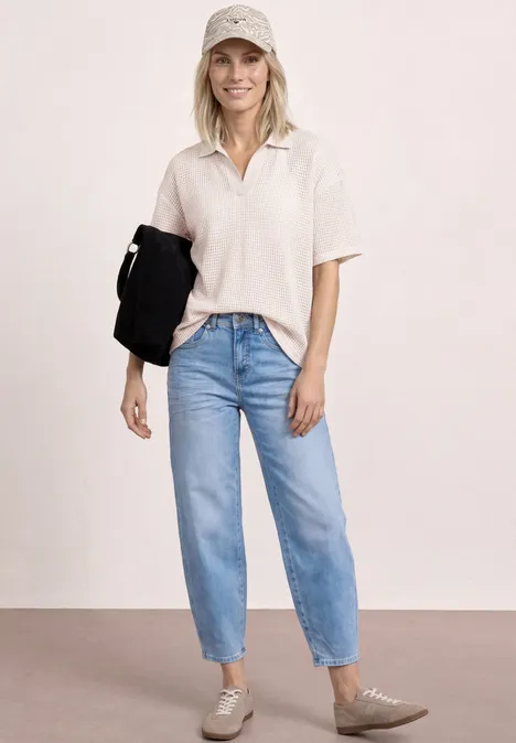 2-in-1 mesh poloshirt met korte mouwen clay sand Een slanke vrouw draagt een licht polo, blauwe jeans, beige schoenen en houdt een zwarte jas vast.