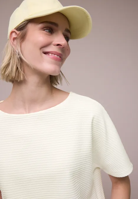 Shirt met structuur in effen kleur off white Vrouw met lichtgele pet, witte gestructureerde top en halflang blond haar.