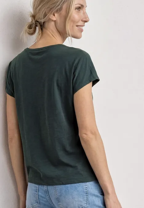 Shirt met decoratief detail deep bound green Blonde vrouw met knot, groen T-shirt en spijkerbroek gezien van achter voor een witte muur